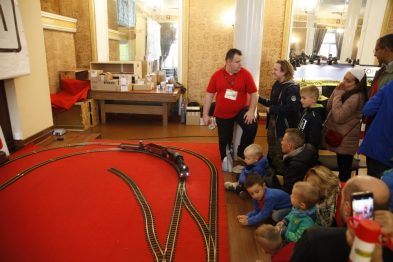 Osoby oglądają makietę kolejową umieszczoną na czerwonym stole. Mężczyzna w koszulce stojący z lewej strony patrzy na dzieci siedzące przed stołem. W tle znajdują się inne osoby i ekspozycje związane z tematem kolejnictwa.