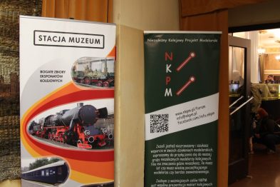 Dwa roll-up bannery stoją obok siebie na ciemnym korytarzu; jeden z nich prezentuje informacje o 