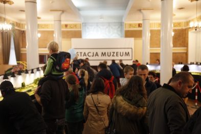 Grupa osób stoi wokół makiety kolejowej, obserwując jej detale. W tle widoczny jest biały napis „STACJA MUZEUM” umieszczony wysoko na ścianie. W centrum uwagi jest duża makieta z zielonymi obszarami i torami kolejowymi.