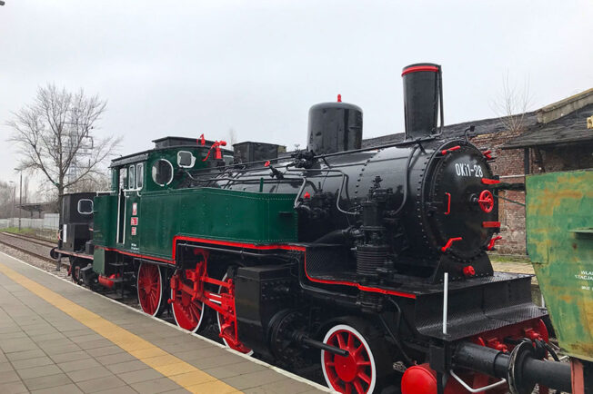 Czarno-zielona lokomotywa parowa z czerwonymi elementami stoi na torach obok peronu. Lokomotywa ma na boku numer 