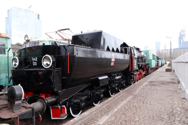 Czarno-czerwona lokomotywa parowa z białymi obramówkami stoi na torach wzdłuż peronu; na boku widoczny jest numer 