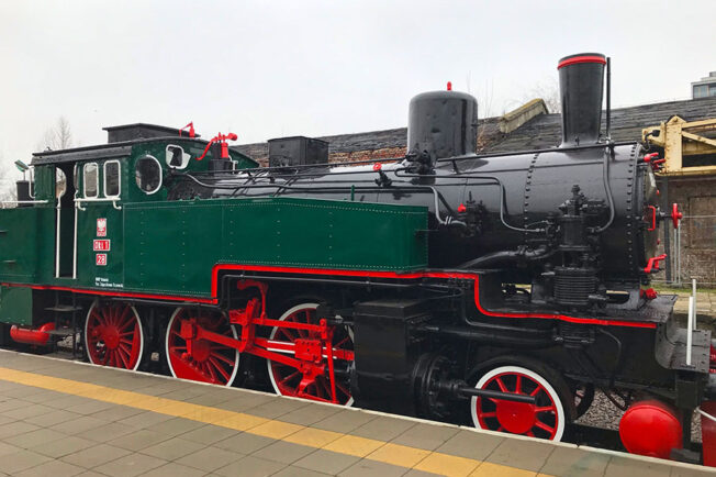 Lokomotywa parowa stoi na torach, jest pomalowana na zielono z czerwonymi elementami i detalami. Na boku widoczny jest czarny numer '28' oraz czerwone obramowania na kołach. Maszyna posiada charakterystyczne atrybuty lokomotyw parowych, takie jak duży kocioł, kominy oraz złożone mechanizmy napędowe.