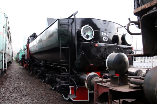 Zdjęcie przedstawia czarną, zabytkową lokomotywę parową oznaczoną numerem Ty42-120, która stoi na torach kolejowych. Cechą charakterystyczną są duże koła napędowe, a także okrągłe okna w kabinie maszynisty. Przed lokomotywą zamontowano czerwone zderzaki oraz znajduje się też czujnik, który jest częścią sprzętu kolejowego.