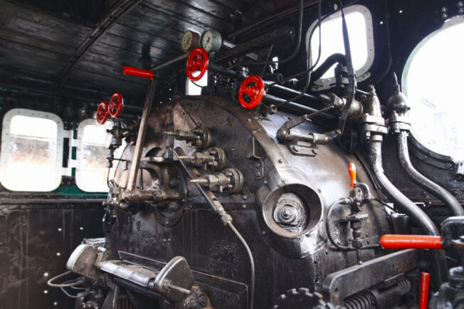 Część wewnętrzna kabiny lokomotywy parowej ukazująca kontrolne tablice, wskaźniki i różnorodne dźwignie. Metalowe powierzchnie charakteryzują się połyskiem, świadcząc o dobrej konserwacji i czystości. Widoczne są zaokrąglone okna kabiny, które zapewniają oświetlenie i widoczność dla obsługi.