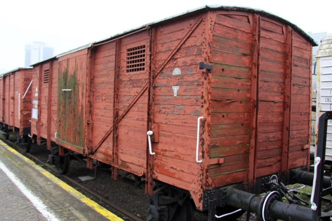 Dwuosiowy wagon kryty o konstrukcji drewnianej prezentuje się na tle peronu, z widocznych drzwiami i okienkami wentylacyjnymi na bokach. Jego czerwona farba jest wyblakła, a białe elementy oznaczeń kontrastują z ogólnym wyglądem. Wózek towarowy jest częścią składu kolejowego, zaparkowanego na szynach.