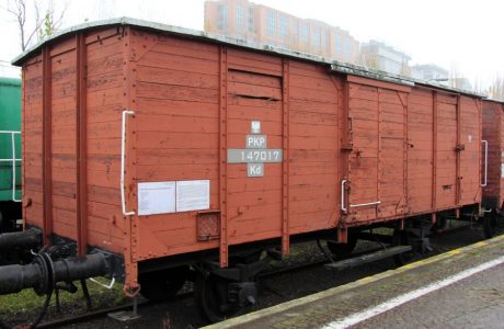 Czerwony, dwuosiowy wagon kryty stoi na torach kolejowych. Konstrukcja wagonu jest drewniana z widocznymi metalowymi elementami jak okucia i zaczepy drzwi, a na bocznej ścianie znajduje się białe oznaczenie przewoźnika i numer seryjny. Dźwi wagonu są zamknięte i zakotwiczone.