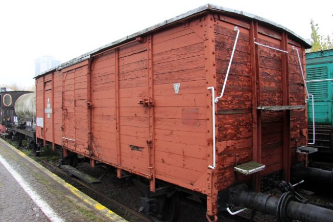 Dwuosiowy wagon towarowy o konstrukcji drewnianej stoi na torach kolejowych. Wagon posiada metalowe elementy wzmacniające, drzwi przesuwne i poziome deski na bokach. Znajduje się na zewnątrz, a tło stanowią inne pojazdy szynowe oraz drzewa.