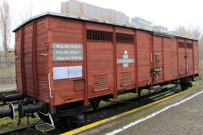 Stary wagon kolejowy stoi na torach; jest to model dwuosiowy z drewnianymi ścianami i metalowymi elementami. Posiada kilka dużych, przesuwnych drzwi i jest pomalowany na czerwono. W tle widoczne są fragmenty budynków i drzewa.