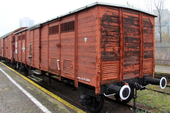 Drewniany wagon towarowy stoi na torach kolejowych; malowany na czerwono, z widocznymi drzwiami i klamkami. Wagon posiadający dwie osie i czarne, prostokątne okna wentylacyjne, jest charakterystyczny dla starszych modeli. Po bokach wagonu znajdują się białe napisy z informacjami.