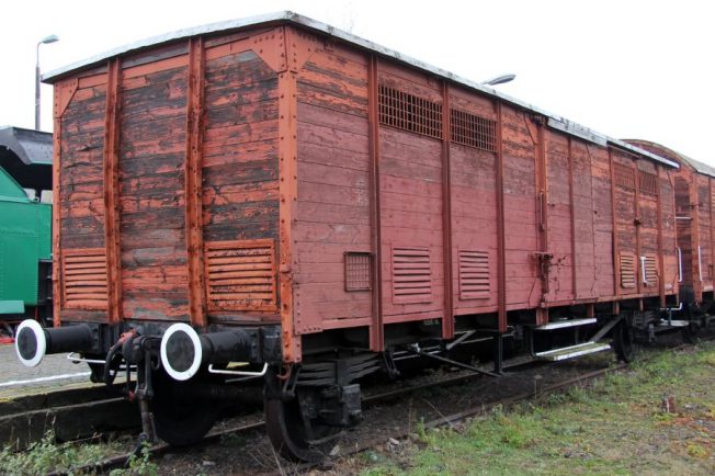 Stary, czerwony wagon towarowy stoi na torach kolejowych, otoczony lekką warstwą trawy i nieco gruntu. Wagon posiada drewniane elementy na bocznych ścianach i białe odbijacze przy końcach. Na środku przedniej ściany widoczne są drzwi, a po bokach kraty wentylacyjne.