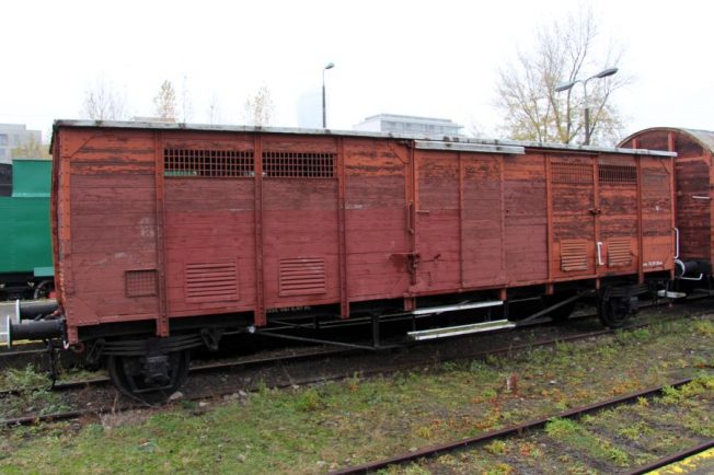 Czerwony dwuosiowy wagon towarowy stoi na torach; ma wiele drzwi z żaluzjami i poszycie. Konstrukcja wagonu wskazuje na starszy model z początku XX wieku. Wagon jest częścią ekspozycji kolejowej, otoczony innym sprzętem kolejowym.