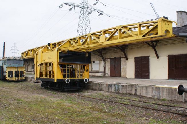 Żółty dźwig dwuramienny stoi na trzyosiowej platformie kolejowej o numerze 856326 przy peronie stacji. Wysięgnik o długości 24 metrów wskazuje na jego zdolność do manipulacji ciężkimi elementami torowymi. Obok znajduje się inny, starszy pojazd kolejowy w odcieniach niebieskiego i żółtego.