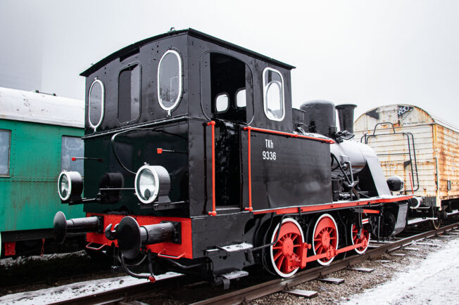 Czarno-czerwona lokomotywa parowa z okrągłym przodem stoi na torach kolejowych; numery 