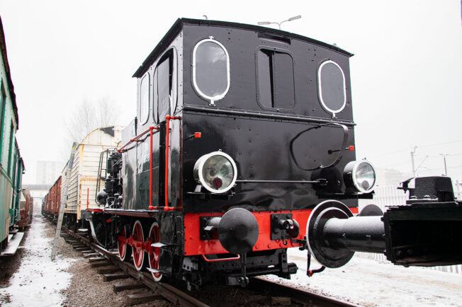 Czarna lokomotywa parowa z czerwonymi obramówkami stoi na torach. Posiada okrągłe białe reflektory i wielkie, okrągłe okna kabiny. Za parowozem widoczny jest ciąg wagonów towarowych.