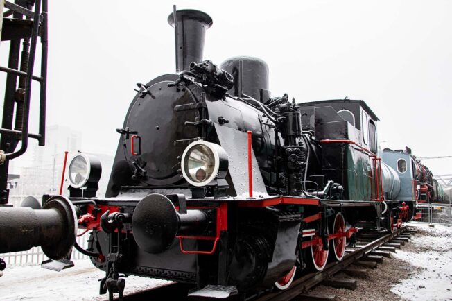 Czarna lokomotywa parowa z czerwonymi obramowaniami stoi na torach; ma duży, okrągły komin i charakterystyczną, okrągłą latarnię z przodu. Obok lokomotywy znajduje się ciemnozielony wagon, również z czerwonymi elementami. Na zewnątrz widać lekki śnieg pokrywający ziemię i częściowo tory.
