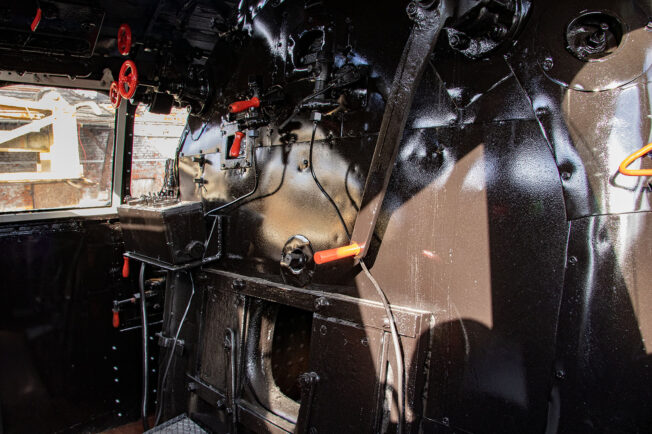 Wewnątrz ciemnej kabiny lokomotywy widoczne są różne przyrządy i wskaźniki o metalicznych, lśniących powierzchniach, z czerwoną oznakowaniem i pokrętłami. Po bokach kabiny przebiegają ciemne rury, a na ścianach i sufit wyraźnie odznaczają się elementy konstrukcyjne. W centrum zainteresowania jest świecąca żarówka o pomarańczowym świetle, która rzuca blask na pobliskie elementy.