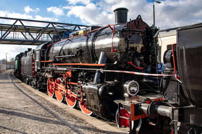 Czarno-czerwona lokomotywa parowa oznaczona numerem Tr203-451 stoi na torach kolejowych. Posiada wiele rur, kół i komponentów typowych dla tradycyjnych parowozów, w tym duży zbiornik na wodę i cylindry napędowe. Lokomotywa jest starannie odrestaurowana i prezentuje się majestatycznie pod jasnym niebem; w tle widoczna jest metalowa kładka dla pieszych.