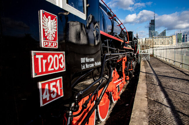 Masywna, czarna lokomotywa parowa z czerwonymi akcentami stoi na torach, a słońce zaświeca na bok pojezdu, wydobywając szczegóły konstrukcji. Umieszczone znaki, w tym biało-czerwony herb, sugerują, że pociąg jest własnością polskich kolei państwowych, a numer 451 widoczny jest na czerwonym tle. W tle widoczne są elementy nowoczesnej infrastruktury, w tym wysokie budynki i częściowo niebieskie niebo