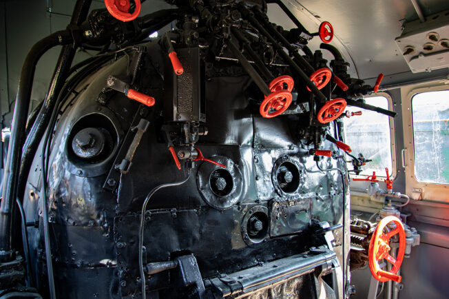W kabinie maszynisty lokomotywy parowej widoczne są skomplikowane mechanizmy sterujące. Na pierwszym planie wyróżniają się czerwone uchwyty i manometry, a także liczne zawory i pokrętła. Panele i ściany kabiny pokryte są różnego rodzaju przyrządami kontrolno-pomiarowymi.