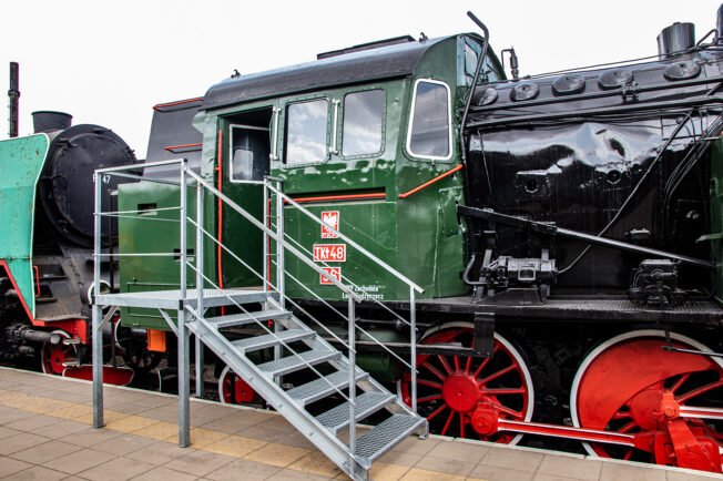 Czarna lokomotywa parowa z czerwonymi kołami i elementami, oznaczona numerem TKt48-36, stoi na torach; obok znajduje się metalowa, szara drabina. Na ścianie lokomotywy widać emblemat z oznaczeniem i białą gwiazdą. Lokomotywa jest w dobrym stanie z lśniącą farbą i wygląda na zadbana.