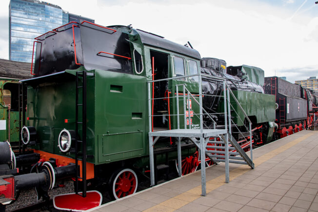 Lokomotywa parowa o zielono-czarnym malowaniu stoi na torach kolejowych obok peronu. Posiada czerwone elementy na kołach oraz białe obramowania i napisy. Wyposażona jest w schodki prowadzące do kabiny maszynisty i osłony na koła; zaczepiona jest do innych wagonów lub lokomotyw.