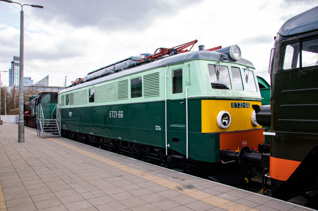 Zielona lokomotywa elektryczna o oznaczeniu ET21-66 stoi na torach; jest ona model z serii produkowanej między 1957 a 1971 rokiem. Maszyna posiada charakterystyczny wygląd z tamtego okresu z wyraźnymi, żółtymi akcentami na przednim zderzaku i bokach. Lokomotywa jest zatrzymana obok peronu i innych składów kolejowych w tle.