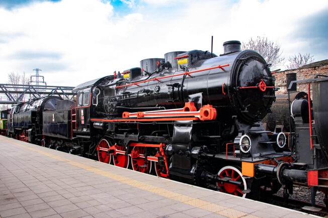 Czarna lokomotywa parowa z czerwonymi obramowaniami kół i elementów znajduje się na peronie obok płaskiej platformy z niebieskim niebem w tle. Maszyna ma duży czarny kocioł, długi dymnik i komorę na węgiel z tyłu, oraz białe napisy i oznaczenia. Starszy model lokomotywy prezentuje się w dobrym stanie, będąc eksponatem kolejnictwa lub czynną atrakcją turystyczną.