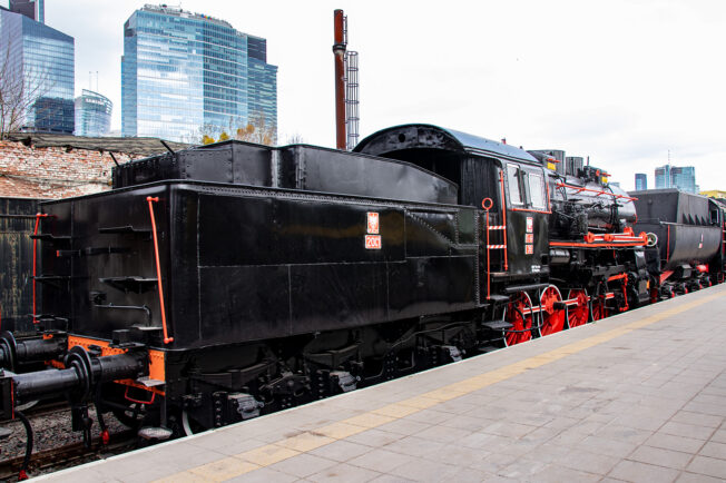 Czarna lokomotywa parowa z czerwonymi elementami jest zaparkowana na torach kolejowych. W tle widoczne są nowoczesne budynki z dużymi oknami i stalową konstrukcją. Przed lokomotywą stoi czarny tender, który służył do przechowywania paliwa i wody.