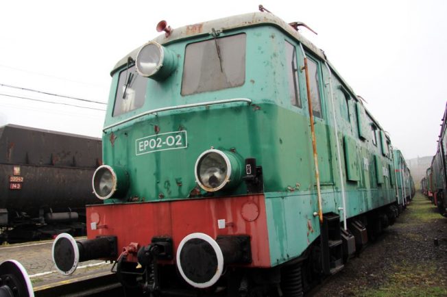 Lokomotywa elektryczna o zielono-czerwonej kolorystyce stoi na torach kolejowych, za nią znajduje się ciemniejszy wagon towarowy. Pojazd jest wyposażony w reflektory, pantografy na dachu oraz wał sprzęgu na przedzie. Maszyna nosi oznaczenie 