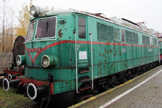 Sześcioosiowa zielona lokomotywa elektryczna z czerwonym pasem stoi na torach. Model posiada liczne okna, drzwi oraz wyraźne oznaczenie EU20-24 na boku. To jedna z lokomotyw dostarczonych dla PKP w latach 50-tych, stojąca na zewnątrz, prawdopodobnie jako eksponat.
