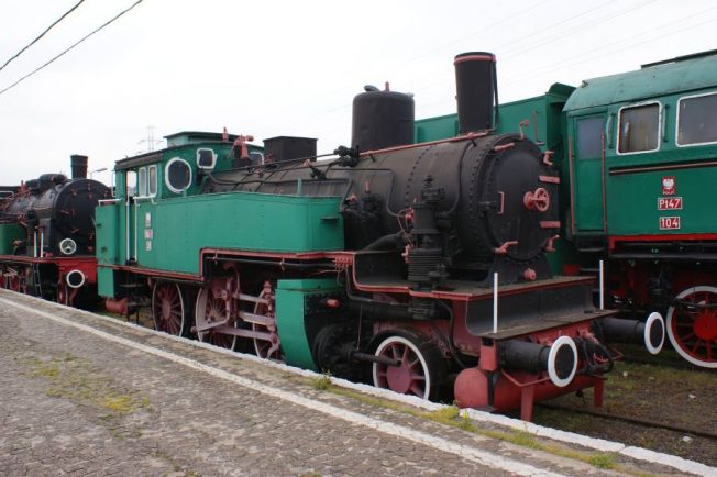 Zielona lokomotywa parowa stoi na torach kolejowych obok innych pojazdów szynowych. Posiada ona czarny kocioł, czerwone obramowania i jest wyposażona w zestawy kół. Z tyłu widoczna jest część innego zielonego pojazdu kolejowego.