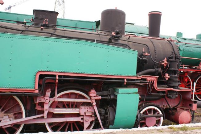 Zabytkowa lokomotywa parowa o klasycznej konfiguracji przedstawia przykład historycznej inżynierii kolejowej. Maszyna ma zielone i czerwone elementy nadwozia ze szczegółami w kolorze czarnym. Duże koła parowozu oraz charakterystyczne elementy jak dymnice i kotły są widoczne i wskazują na technologię z początków XX wieku.