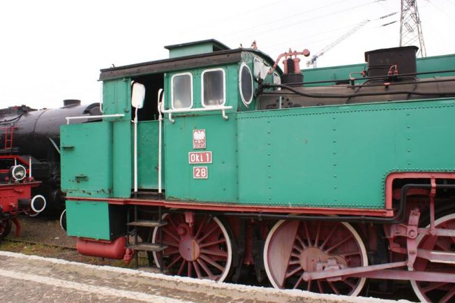 Lokomotywa parowa o oznaczeniu OKi1-28 ma zielone malowanie z czerwonymi kołami i białymi obręczami. Posiada boczną kabinę z białymi oknami, schodki wejściowe i kompaktową budowę. Lokomotywa eksponowana jest na torach, a tuż za nią widać część innej lokomotywy.