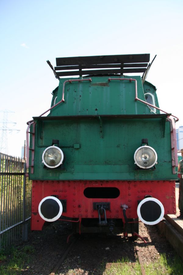 Stara zielona lokomotywa parowa z czerwonymi elementami stoi na torach. Posiada charakterystyczne duże okrągłe reflektory na przodzie oraz liczne elementy metalowe i rury. Wóz wyposażony jest w schodki i poręcze ułatwiające dostęp do kabiny maszynisty.