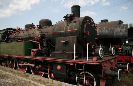 The classic design steam locomotive stands on the tracks; the engine has steering wheels at the front and a set of larger driving wheels. The black locomotive, with red and white markings, has a firebox, a chimney, and a system of pipes and tanks around the engineer's cabin. There are also side panels with numbering and circular, hinged covers on the side steam tanks.