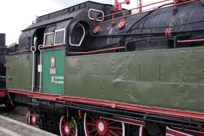 Ciężka lokomotywa parowa o zielonym lakierze stoi na torach kolejowych. Ma charakterystyczne czerwone obręcze kół i komplet ciemnych rur oraz zbiorników, wśród których znajduje się duży, czarny komin. Po jej boku znajdują się nieduże okrągłe okna i tabliczka z informacjami.