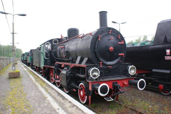 Czarno-czerwona lokomotywa parowa stoi na torach kolejowych. Za nią widoczne są inne wagony i lokomotywy ustawione w szeregu. Lokomotywa ma okrągłe lampy, duże koła oraz jest wyposażona w parę przegrzaną.