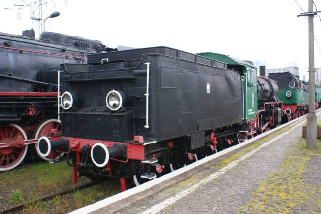 Czarna lokomotywa parowa z czerwonymi elementami takimi jak koła i odbojniki stoi na torach kolejowych. Tuż za nią widoczna jest inna zielona lokomotywa. Obie maszyny są dobrze zachowane, znajdują się na na zewnątrz w dzień.