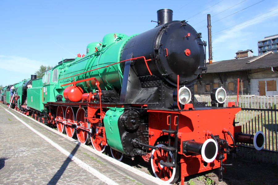 Zielona lokomotywa parowa z czerwonymi elementami stoi na torach kolejowych pod błękitnym niebem. Maszyna, oznaczona numerem Os24-7, posiada czarny, okrągły komin i duże, czarne koła. Na jej bokach widoczne są okrągłe, czarne pokrywy oraz czerwone i białe szprychy kół, a przednia część jest wyposażona w dwie pary okrągłych reflektorów i charakterystyczny, czerwony zderzak.