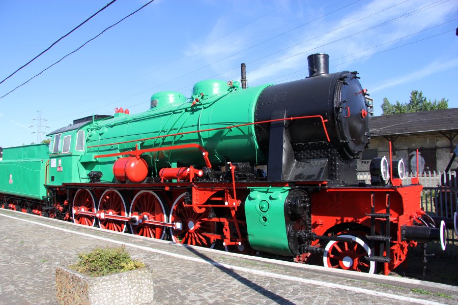 Zielona lokomotywa parowa z czerwonymi kołami stoi na torach w słoneczny dzień. Połączona jest z zielonym tendrem, który prawdopodobnie służy do przechowywania węgla i wody. Obiekty w tle sugerują, że lokomotywa znajduje się na stacji czy muzeum kolejnictwa.