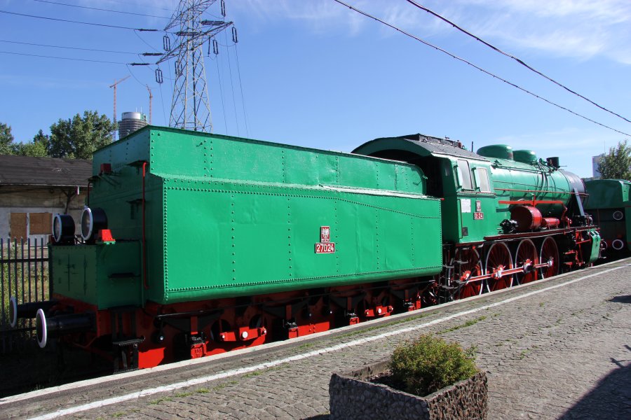 Zielona lokomotywa parowa z czerwonymi obręczami na kołach stoi na torach. Maszyna ma białe linie akcentujące krawędzie i numery identyfikacyjne. Kocioł parowy i klasyczne kształty konstrukcyjne wskazują na historyczny charakter pojazdu.