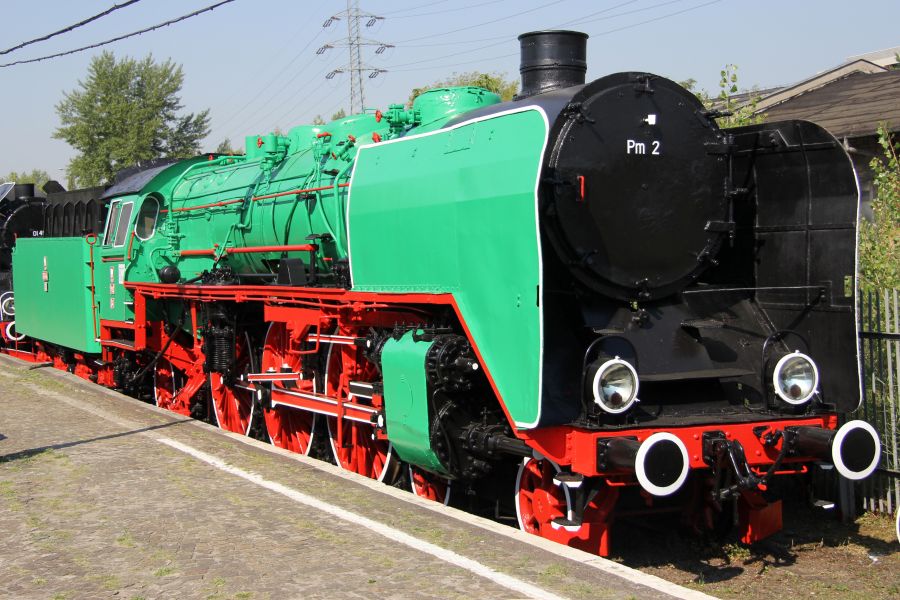 Lokomotywa parowa o zielono-czarnej kolorystyce stoi na torach kolejowych. Maszyna ma duże, czerwone koła i jest wyposażona w wielki czarny kocioł. Na bocznej ścianie lokomotywy widnieje numer Pm2-34.