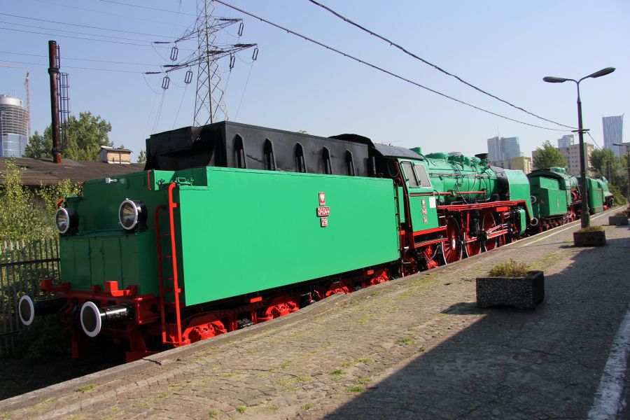 Lokomotywa parowa z zielonym kotłem i czarnym podwoziem stoi na torach kolejowych. W tle zarysowują się drzewa i słupy elektryczne pod niebieskim niebem. Po bokach lokomotywy widoczne są czerwone elementy, takie jak koła i ramy boczne.