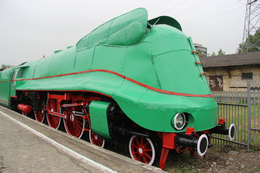 Zachowana w dobrym stanie zielona lokomotywa parowa z czerwonymi detalami stoi na torze kolejowym. Posiada charakterystyczną aerodynamiczną osłonę w kształcie stożka z przodu, a jej duże okrągłe reflektory są wyraźnie widoczne. Kółka lokomotywy i jej elementy są pomalowane na czerwono, co kontrastuje z głównym kolorem lokomotywy.