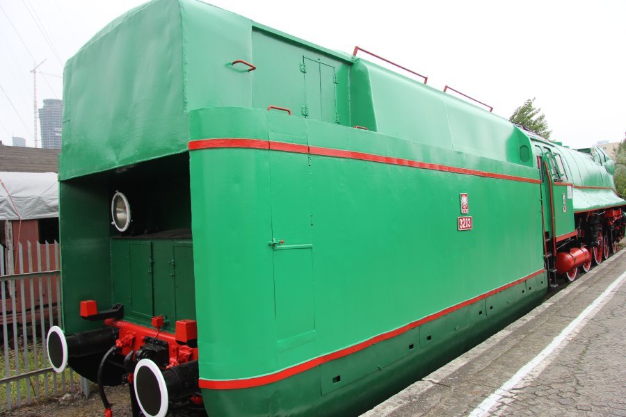 Zielona lokomotywa parowa z czerwonymi obramówkami stoi na torach kolejowych. Posiada ona aerodynamiczną obudowę, dużą osłonę przedniej części i okrągłe okno na bocznej ścianie. Koła i elementy konstrukcji są pomalowane na czarno, a na boku widać mały napis i emblemat.