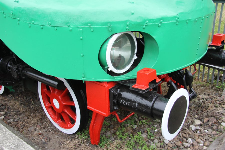 Widoczna jest część zielono pomalowanej lokomotywy parowej z czerwoną obręczą kół i czarnymi elementami. Na pierwszym planie znajduje się bufor oraz cylindryczne elementy, a w tle widać fragment toru kolejowego i kamienie. Okrągły reflektor umieszczony jest na przedzie lokomotywy, a wszystkie części wydają się być w dobrym stanie.