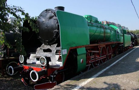 The green and black steam locomotive from the post-war period is on display outside on the railroad tracks. The central part of the heavy vehicle is equipped with numerous complex mechanical elements, including a distinctive large steam boiler and cylinders. The locomotive has red wheel rims and a black, rounded front part with a headlight.