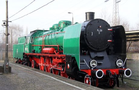 The green and red steam locomotive stands on the tracks; its large wheels and cylindrical front give it a classic look. The black, round cover on the front appears to be freshly painted. The locomotive is surrounded by railway installations, and in the background, trees and traction poles are visible.