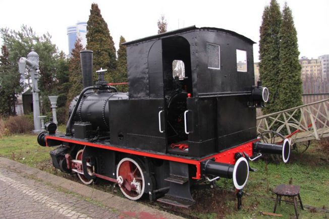 Czarna lokomotywa parowa o prostej konstrukcji z białymi obrzeżami stoi na torach. Konstrukcja obejmuje cylindryczny kocioł, kabina maszynisty jest umieszczona z tyłu. Pojazd jest dwuosiowy z czerwonymi kołami i posiada elementy charakterystyczne dla parowozów manewrowych.