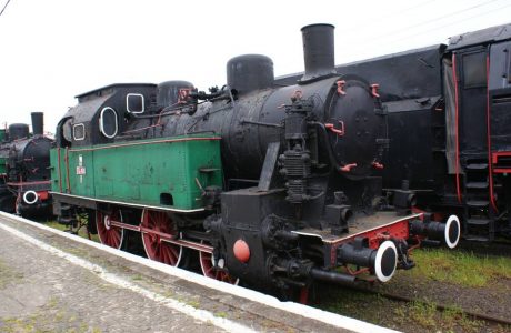 The old steam locomotive marked with the number TKc100-1 stands on the railroad tracks, behind it other wagons or locomotives are visible. The vehicle has a black cab and a green tender, it is equipped with large, red wheels and a cylindrical boiler. Despite its age, the locomotive looks like a well-maintained exhibit from the historical railway era.